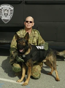 A man in fatigues and a dog sit next to a police vehicle.